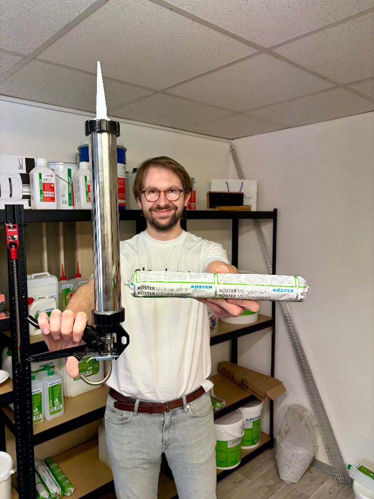 Muž drží v rukou velké 600ml balení tmelu KÖSTER MS-Joint Sealant a profesionální hliníkovou výtlačnou pistoli v zázemí skladu.