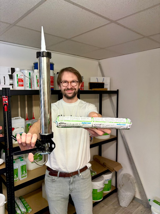 Muž drží v rukou velké 600ml balení tmelu KÖSTER MS-Joint Sealant a profesionální hliníkovou výtlačnou pistoli v zázemí skladu.